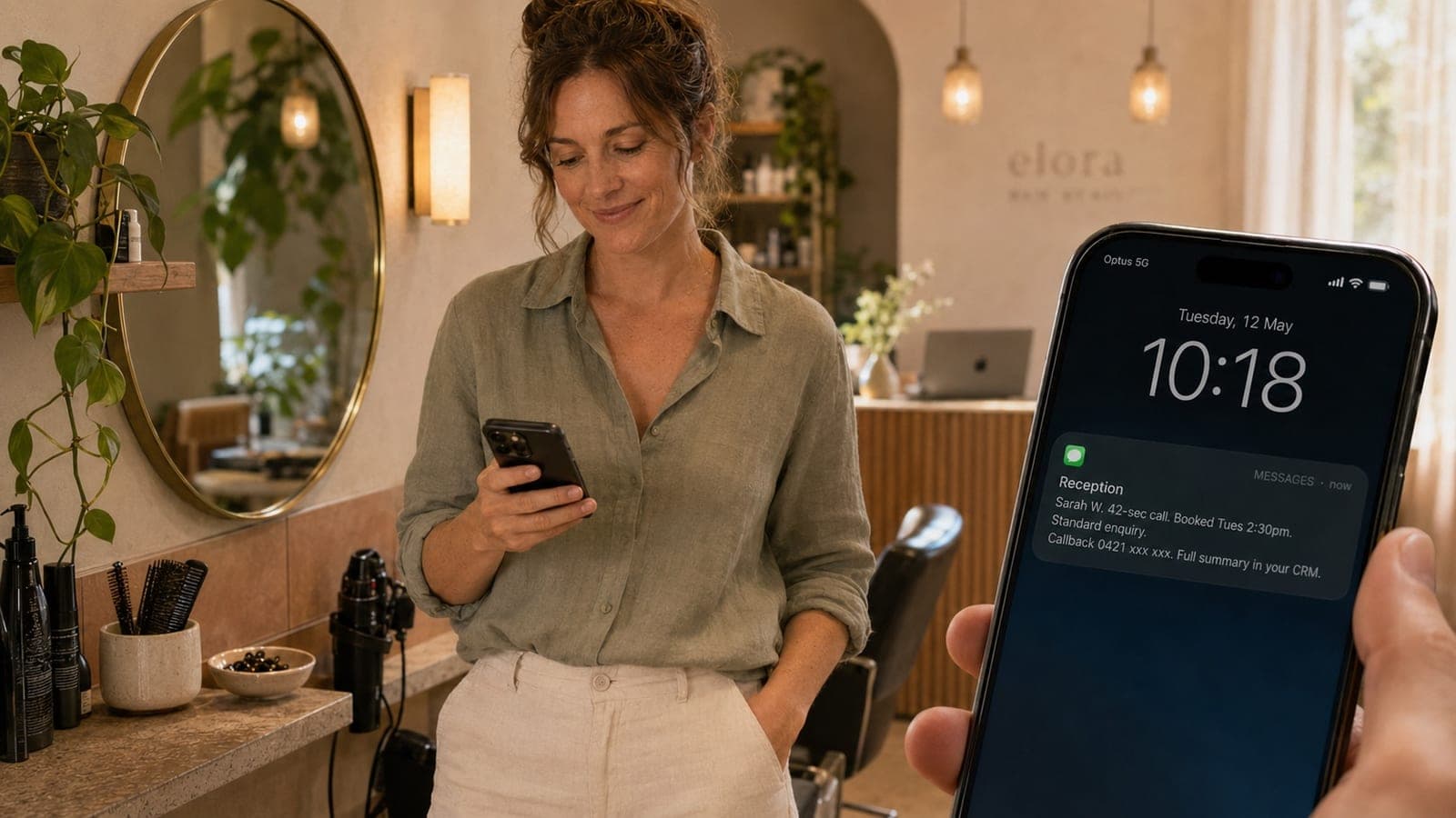 An Australian beauty salon owner standing at her front-of-house, holding her phone and glancing at a text summary from her AI receptionist. A new appointment has just been booked. Soft morning light through sheer curtains, styling station and plants in the background.